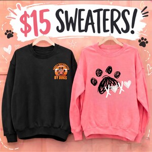 Op de afbeelding: Twee sweatshirts op hangers. Een zwart sweatshirt met de tekst "Easily Distracted By Dogs" en een hondenpoot-afbeelding. De andere is roze met een pootafdruk en hartjes.