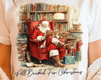May include: White t-shirt featuring a graphic of Santa Claus reading in a library, with the text "All Booked For Christmas" in a script font. Santa is seated in a red armchair surrounded by books. The shirt is a festive holiday apparel item.