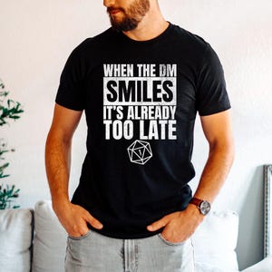 May include: Black t-shirt featuring the text "WHEN THE DM SMILES IT'S ALREADY TOO LATE" in white, along with a white D20 die graphic. The t-shirt has a crew neck.