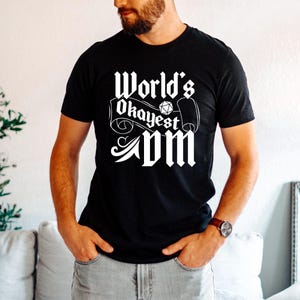 May include: Black t-shirt with the text "World's Okayest DM" in white, gothic-style lettering. The design includes a die and scroll elements. The shirt is a crew neck and is being worn.