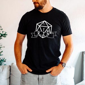 May include: Black t-shirt featuring a white graphic of a D20 die with the number 1, a skull, and the letters "R.I.P." on a banner. The shirt is a short-sleeved crew neck.