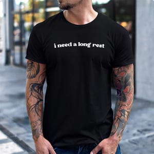 May include: Black crew neck t-shirt with the white text "i need a long rest" across the chest. The shirt is plain and simple, suitable for casual wear. The person wearing the shirt has tattoos on both arms.