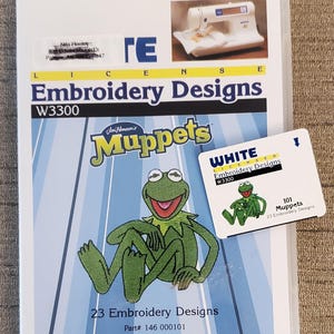 May include: A white plastic case with a blue and white label that reads "Embroidery Designs W3300 Jim Henson's Muppets 23 Embroidery Designs Part# 146 000101". The case contains a white plastic card with a blue and white label that reads "White Licensed Embroidery Designs W3300 101 Muppets 23 Embroidery Designs". The card features a green cartoon frog.