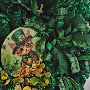 May include: A St. Patrick's Day wreath with a round centerpiece featuring a kitten wearing a green hat and bow tie, sitting on gold coins. The wreath is made of green ribbons and mesh.