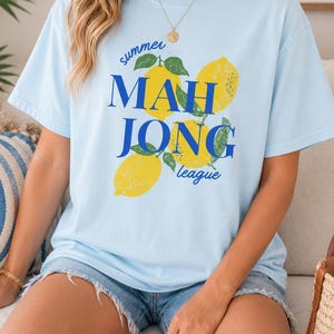 May include: A light blue short-sleeve t-shirt featuring a graphic print of three yellow lemons with green leaves. The text "summer" is printed in small blue script above the words "MAH JONG" in large blue letters, with "league" below.