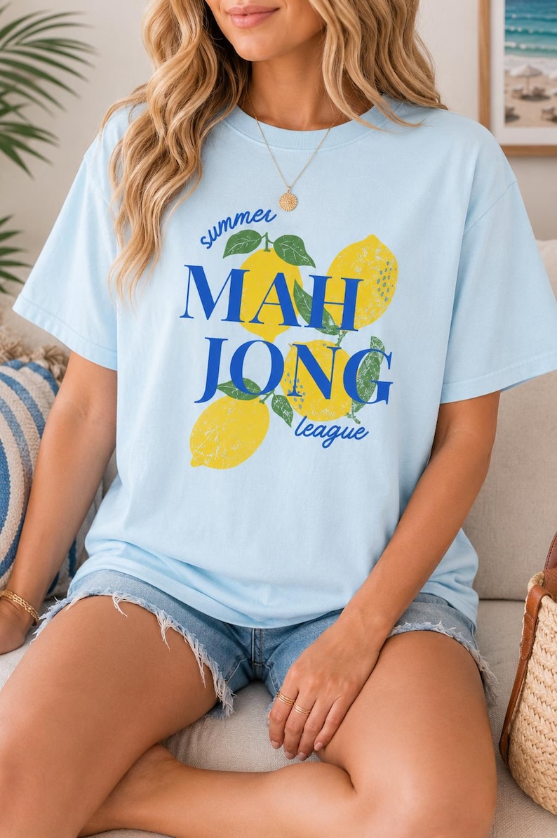 May include: A light blue short-sleeve t-shirt featuring a graphic print of three yellow lemons with green leaves. The text "summer" is printed in small blue script above the words "MAH JONG" in large blue letters, with "league" below.