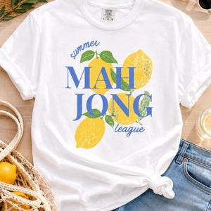 May include: A white short-sleeve t-shirt featuring a graphic print of three yellow lemons with green leaves. The text "summer MAHJONG league" is printed in blue across the center. The shirt has a crew neckline and a Comfort Colors brand label.