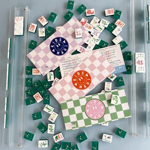 May include: A collection of Kids Mahjong sets with green, pink, and purple checkered boxes. The boxes feature a smiley face logo and the text "KIDS MAH". Scattered around are green and white tiles with various symbols and designs. The sets are displayed on a light blue surface.