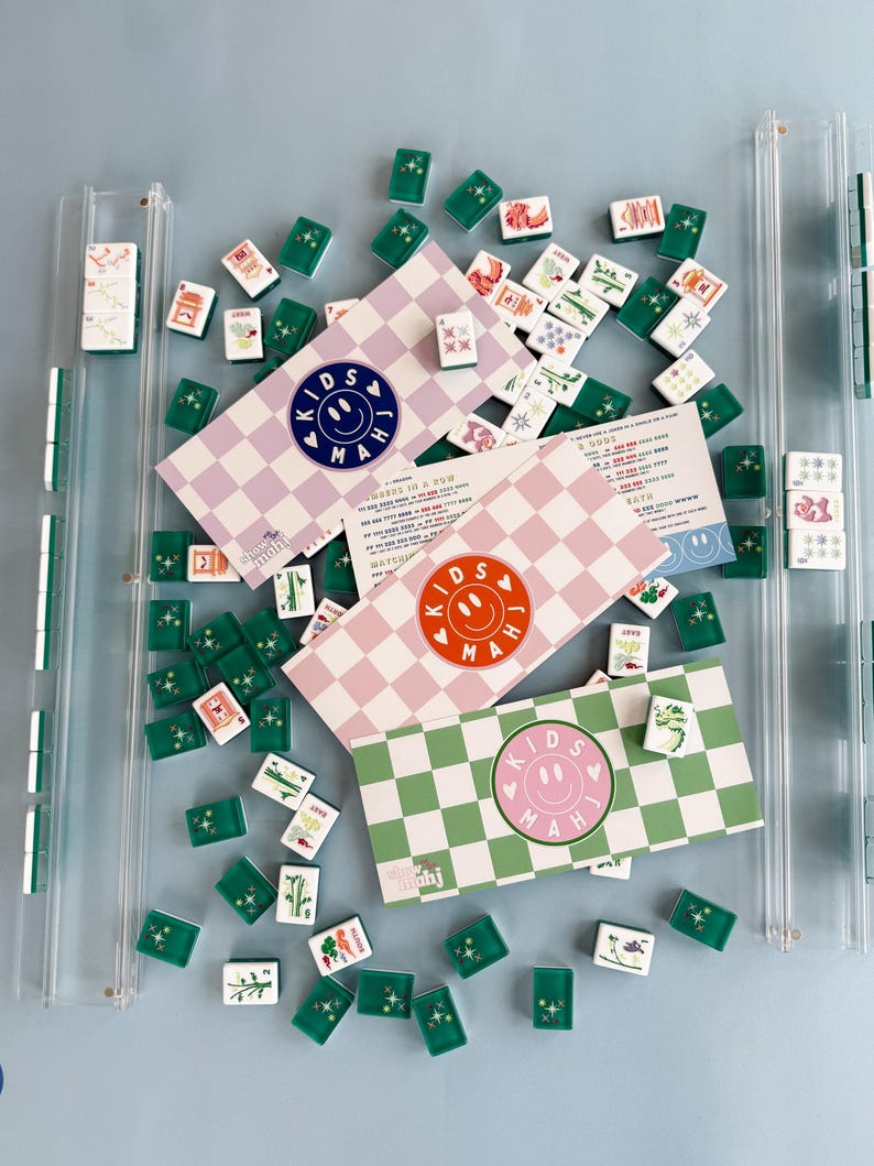 May include: A collection of Kids Mahjong sets with green, pink, and purple checkered boxes. The boxes feature a smiley face logo and the text "KIDS MAH". Scattered around are green and white tiles with various symbols and designs. The sets are displayed on a light blue surface.