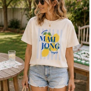 May include: A cream-colored short-sleeve t-shirt featuring a graphic print of yellow lemons and blue vertical stripes. The text reads "summer MAH JONG league" in blue and script lettering. The shirt has a crew neckline and a relaxed fit.