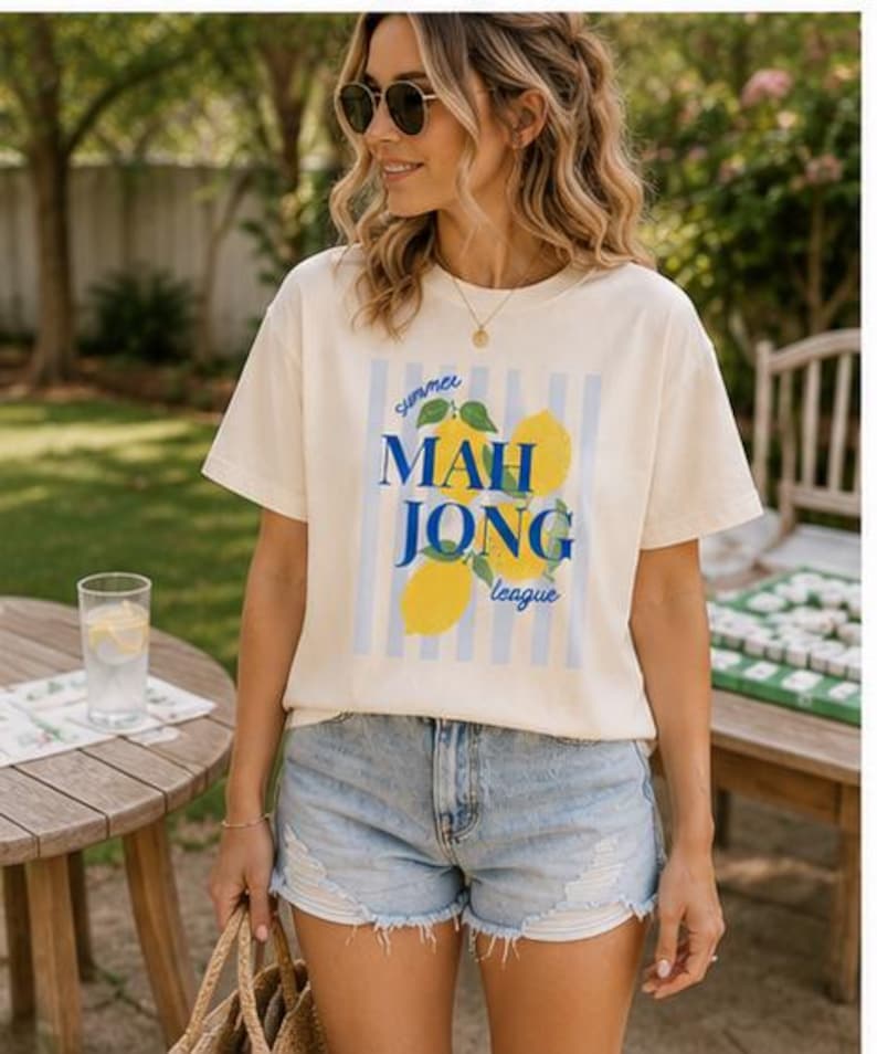 May include: A cream-colored short-sleeve t-shirt featuring a graphic print of yellow lemons and blue vertical stripes. The text reads "summer MAH JONG league" in blue and script lettering. The shirt has a crew neckline and a relaxed fit.