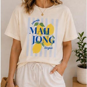 May include: A cream-colored short-sleeve t-shirt featuring a graphic print of yellow lemons with green leaves over light blue vertical stripes. The text reads "summer MAH JONG league" in blue lettering. The shirt has a crew neckline and a relaxed fit.