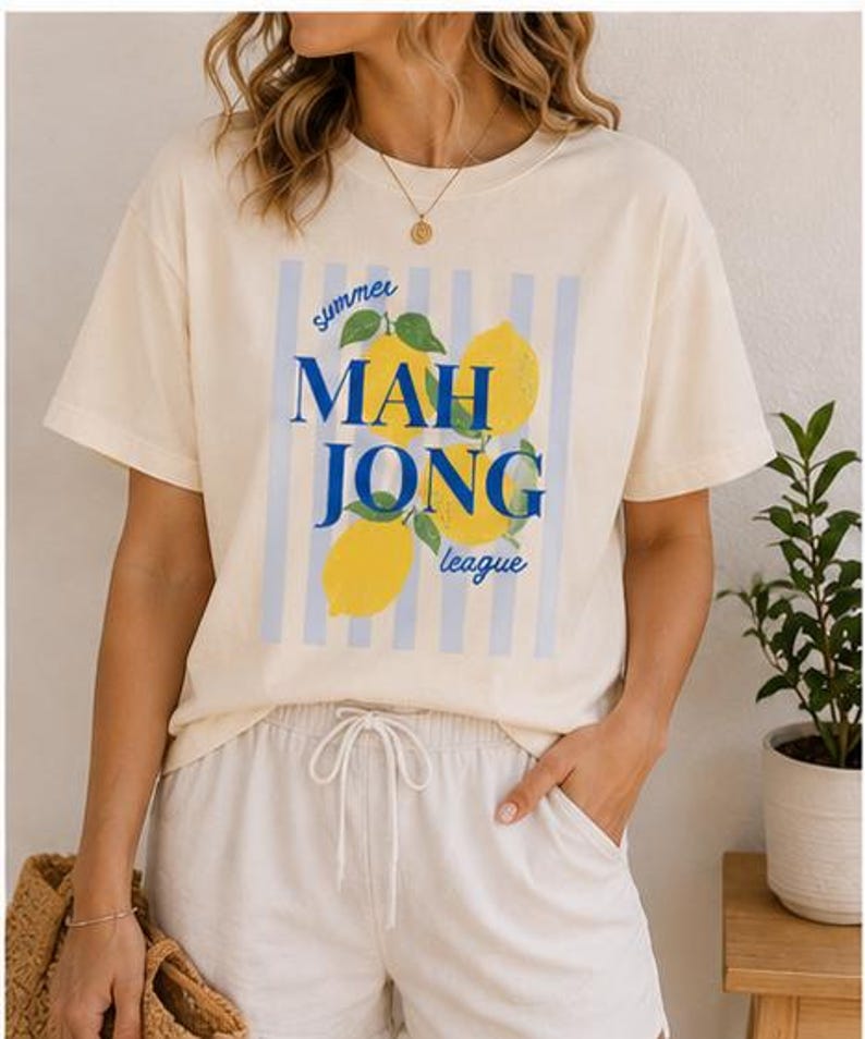 May include: A cream-colored short-sleeve t-shirt featuring a graphic print of yellow lemons with green leaves over light blue vertical stripes. The text reads "summer MAH JONG league" in blue lettering. The shirt has a crew neckline and a relaxed fit.