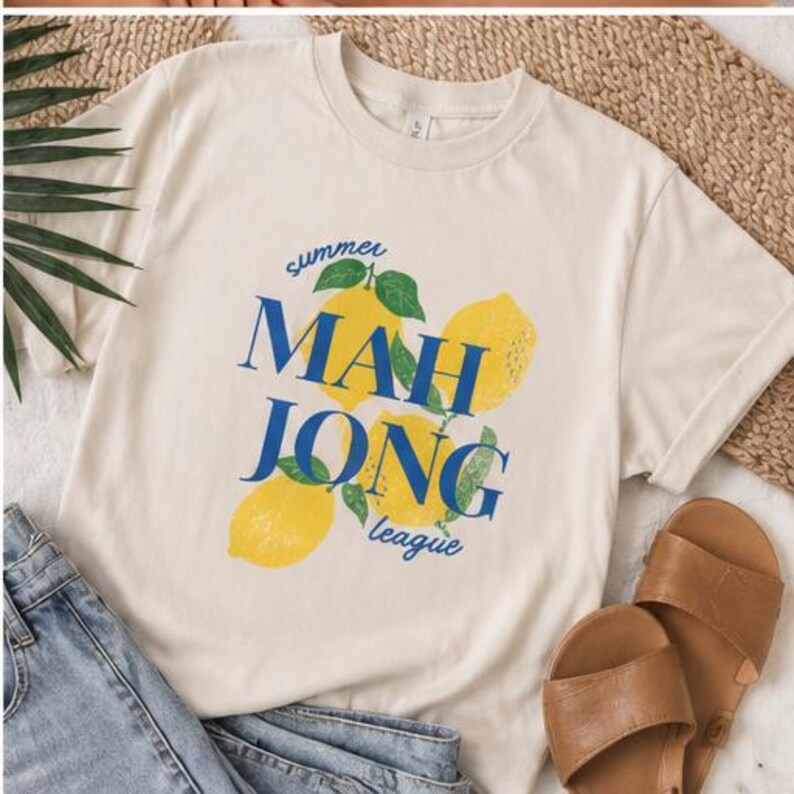 May include: A cream-colored short-sleeve t-shirt featuring a graphic print of three yellow lemons with green leaves. The text "summer" is printed in blue above the lemons, and the blue text "MAH JONG league" is layered over the lemon graphic.