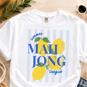 May include: A white short-sleeve t-shirt featuring a graphic print of three yellow lemons with green leaves over a light blue vertical striped background. The text reads summer MAH JONG league in blue, with the Comfort Colors brand label at the neck.