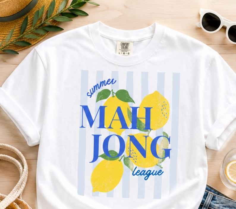 May include: A white short-sleeve t-shirt featuring a graphic print of three yellow lemons with green leaves over a light blue vertical striped background. The text reads summer MAH JONG league in blue, with the Comfort Colors brand label at the neck.