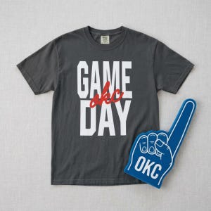 Op de afbeelding: Donkergrijs t-shirt met de woorden "GAME OKC DAY" in witte blokletters en rode cursief. Een blauwe foam finger met "OKC" in wit ligt naast het shirt. Ideaal voor de wedstrijddag.