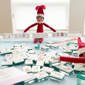 May include: A red-suited elf figure with a red hat and white trim is positioned above a collection of Mahjong tiles. The tiles are white with various colorful symbols and sit on a light blue surface. A second elf figure is visible.