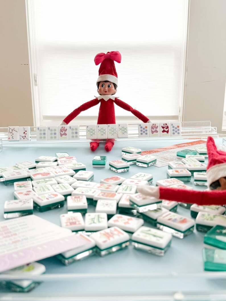 May include: A red-suited elf figure with a red hat and white trim is positioned above a collection of Mahjong tiles. The tiles are white with various colorful symbols and sit on a light blue surface. A second elf figure is visible.