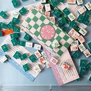 May include: A collection of Mahjong tiles and game materials. The tiles are green and white, with various symbols. The game box has a green and white checkered pattern with the text "KIDS MAHJ" and a smiley face. Several cards with smiley faces and text are scattered around.