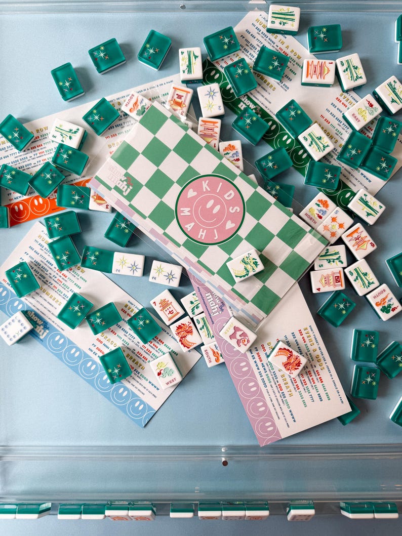 May include: A collection of Mahjong tiles and game materials. The tiles are green and white, with various symbols. The game box has a green and white checkered pattern with the text "KIDS MAHJ" and a smiley face. Several cards with smiley faces and text are scattered around.