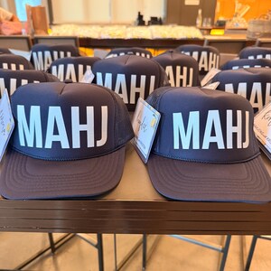 May include: A collection of navy blue trucker hats featuring a mesh back and a structured front panel. Each hat displays the white text MAHJ in a bold, sans-serif font across the front. A small rectangular tag is attached to the side of the hats.