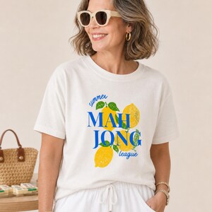 May include: A cream-colored short-sleeve t-shirt featuring a graphic print of yellow lemons with green leaves and the blue text "summer MAH JONG league" centered on the front. The shirt has a crew neckline and a relaxed fit.