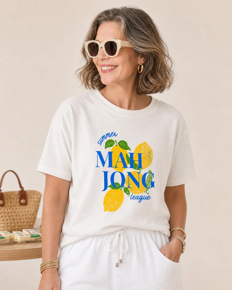 May include: A cream-colored short-sleeve t-shirt featuring a graphic print of yellow lemons with green leaves and the blue text "summer MAH JONG league" centered on the front. The shirt has a crew neckline and a relaxed fit.