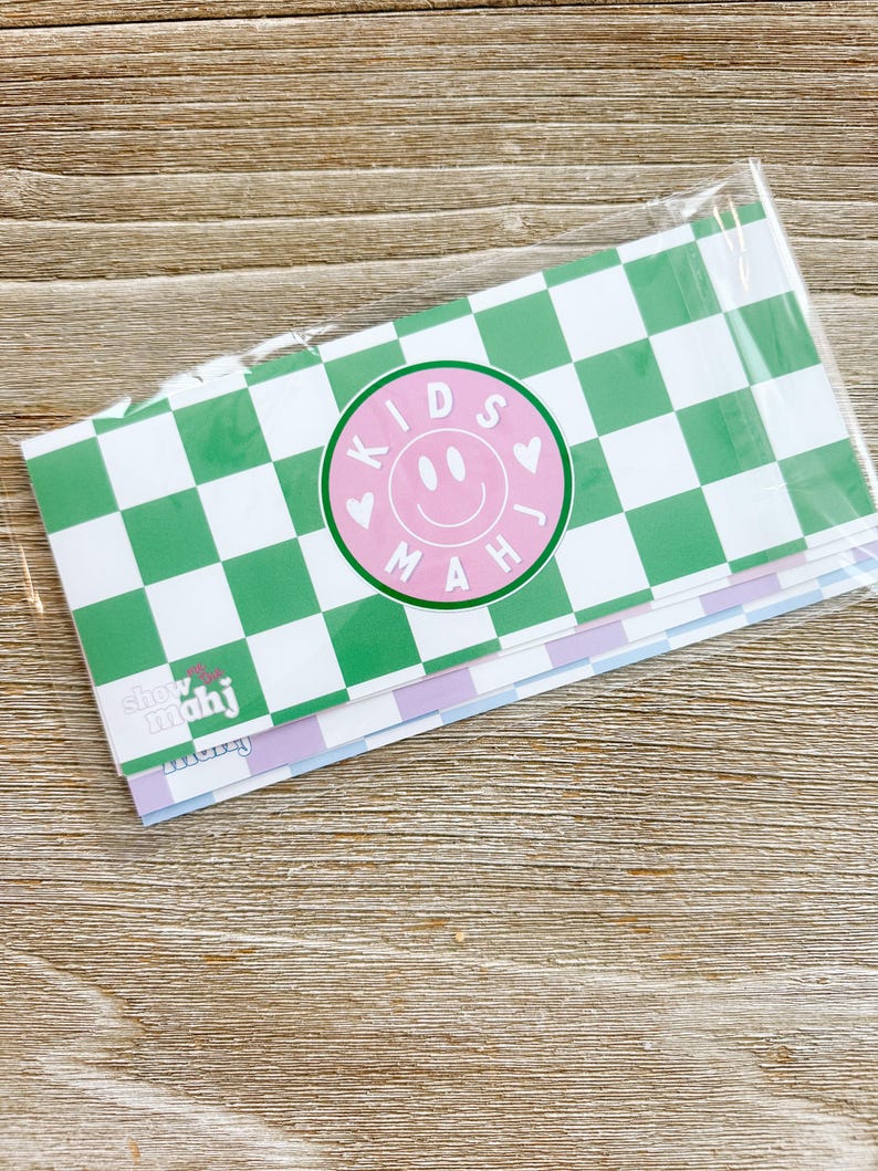 May include: A stack of stationery with a green and white checkered pattern. A pink circle with a smiley face and the words "KIDS MAH" is centered on the top sheet. The text "show mahj" is visible on the bottom sheet.