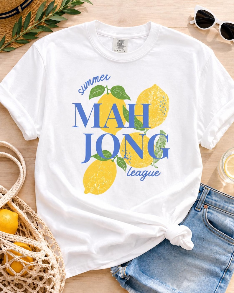 May include: A white short-sleeve t-shirt featuring a graphic of three yellow lemons with green leaves. The text "summer MAHJONG league" is printed in blue, with "MAHJONG" in large capital letters and "summer" and "league" in script.