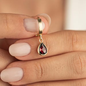 May include: A gold earring featuring a teardrop-shaped, faceted, deep red gemstone pendant. The earring is a small hoop style, and the gemstone is set in a gold bezel. The earring is held between fingers.