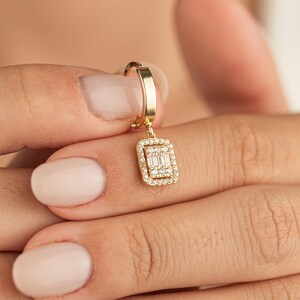 May include: A gold-colored earring with a rectangular pendant. The pendant is encrusted with small, clear gemstones around the edges and features a cluster of baguette-cut stones in the center. The earring is held by a hand with manicured nails.