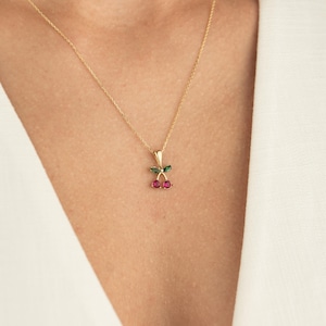 Può includere: Una collana d'oro con un ciondolo a forma di ciliegia. Il ciondolo presenta due pietre preziose rosse che rappresentano le ciliegie, con foglie verdi e uno stelo dorato. La collana è mostrata su uno sfondo bianco.