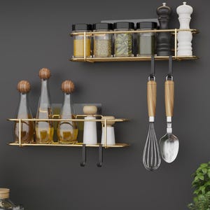 Może przedstawiać: Dwie złote metalowe półki ścienne z haczykami. Górna półka zawiera sześć szklanych słoików na przyprawy z czarnymi pokrywkami i dwie czarne i białe solniczki i pieprzniczki. Dolna półka zawiera trzy szklane butelki z drewnianymi korkami i dwie białe solniczki i pieprzniczki. Dwa drewniane narzędzia kuchenne wiszą na haczykach.