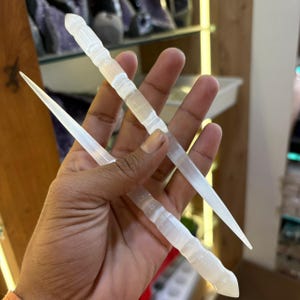 May include: Two white selenite wands are held in a hand. One wand is pointed and the other has a rounded end. The wands have a textured, layered appearance. The background shows shelves with other crystals.