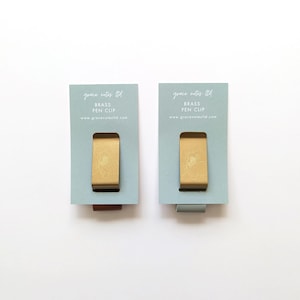 May include: Two brass pen clips with floral designs, displayed on light blue cards. The cards have the text "grace notes ltd", "BRASS PEN CLIP", and a website address. The pen clips are rectangular with a curved top.