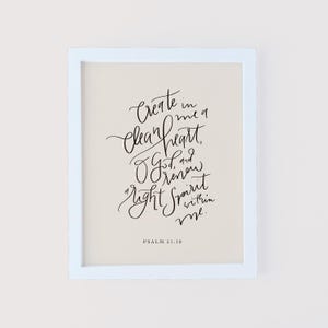 May include: A framed print with a light blue border. The print features elegant black calligraphy on a cream background. The text reads "Create in me a clean heart, O God, and renew a right spirit within me." Psalm 51:10 is written at the bottom.