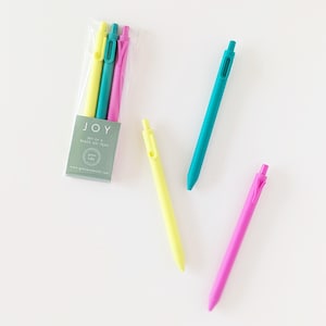May include: A set of black gel pens in a clear plastic package, with the word "JOY" printed on the front. The pens are yellow, teal, and pink. Two additional pens in yellow and pink are also visible.