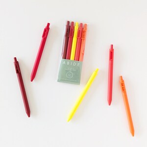 May include: A set of black gel pens in shades of red, orange, and yellow, with the word "ABIDE" printed on the pens and packaging. The pens are scattered on a white surface.
