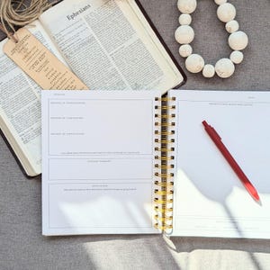 May include: Open Bible with the word "Ephesians" visible, a journal with gold spiral binding, and a red pen. A bookmark with text hangs from the Bible. A white beaded necklace is in the background. The journal has sections for prayers and notes.