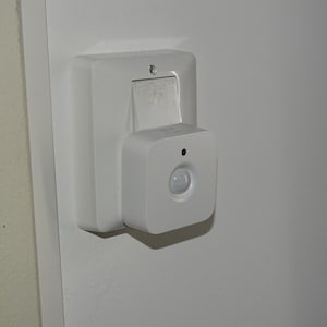 Op de afbeelding: Een witte bewegingssensorarmatuur bevestigd aan een witte muur. De armatuur heeft een rechthoekige basis met een kleinere, vierkante sensoreenheid. De sensor heeft een afgeronde lens en een kleine zwarte stip.