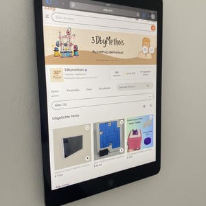 Może przedstawiać: Czarny tablet wyświetlający sklep Etsy 3DbyMethois, z ilustracją drukarki 3D na banerze. Tablet jest zamontowany na białej ścianie, prezentując różne przedmioty.