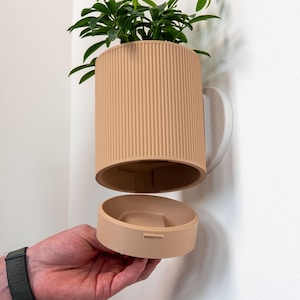 Op de afbeelding: Een aan de muur gemonteerde cilindrische plantenbak met een geribbelde buitenkant in een lichte perzikkleur. De plantenbak heeft een afneembare ronde onderschaal in dezelfde kleur, die los onder de hoofdbehuizing wordt gehouden.