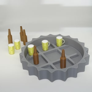 Puede incluir: Una bandeja gris en forma de tapón de botella con botellas de cerveza marrones en miniatura y tazas amarillas con tapas blancas. Varias botellas y tazas se colocan dentro de la bandeja, y otras fuera. Los objetos están dispuestos sobre una superficie blanca.