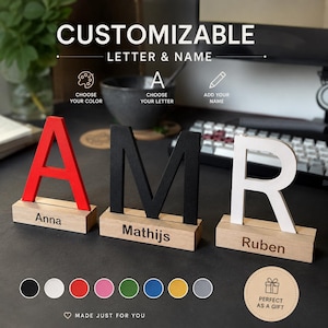 Op de afbeelding: Drie aanpasbare staande letters, A, M en R, in rood, zwart en wit, elk gemonteerd op een rechthoekige houten voet. De voetstukken zijn gegraveerd met de namen Anna, Mathijs en Ruben. Onderaan staan acht gekleurde cirkels.