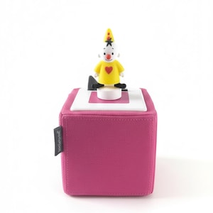 Puede incluir: Un cubo de tela magenta con una parte superior blanca y rosa, con una figura de payaso. El payaso lleva una camisa amarilla con un corazón rojo, un sombrero alto amarillo y zapatos negros. El cubo tiene una pequeña etiqueta negra en el lateral.