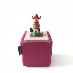 Puede incluir: Un cubo de tela magenta con una parte superior blanca y rosa, con una pequeña figura de una niña con capucha y vestido rojos. La figura está sobre una base verde. El cubo tiene una etiqueta negra que dice "toniebox".