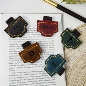Può includere: Set di segnalibri in pelle in vari colori, tra cui nero, marrone, bordeaux e verde acqua. Ogni segnalibro presenta un design unico, come un'impronta di zampa, una lettera, una croce o un nome. I segnalibri sono attaccati a un elastico nero.