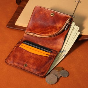 Puede incluir: Cartera de cuero marrón abierta con un bolsillo con cremallera, cierre a presión y múltiples ranuras para tarjetas. La cartera contiene efectivo, tarjetas de crédito y monedas. La cartera está sobre una superficie marrón.