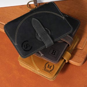 May include: A stack of three rectangular leather wallets in black, dark brown, and tan. Each wallet features a strap closure, visible stitching, and a personalized engraving: the letter S, the letter B, or the letter M with the name MANDY.